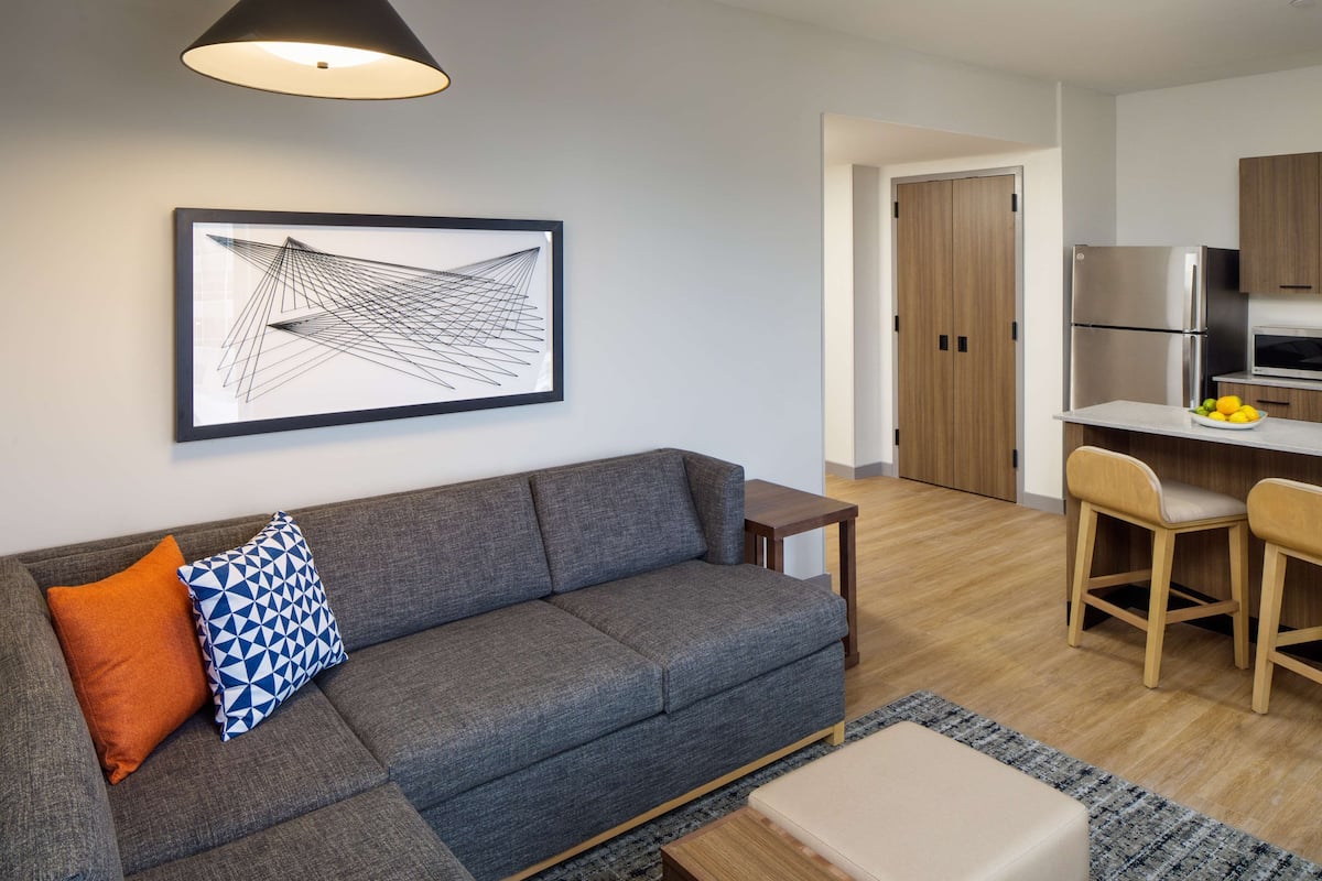 A modern living room with a grey sofa, a coffee table, and a small wooden side table. A framed abstract artwork hangs on the wall, and a kitchen area is visible in the background.