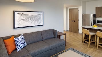 A modern living room with a grey sofa, a coffee table, and a small wooden side table. A framed abstract artwork hangs on the wall, and a kitchen area is visible in the background.