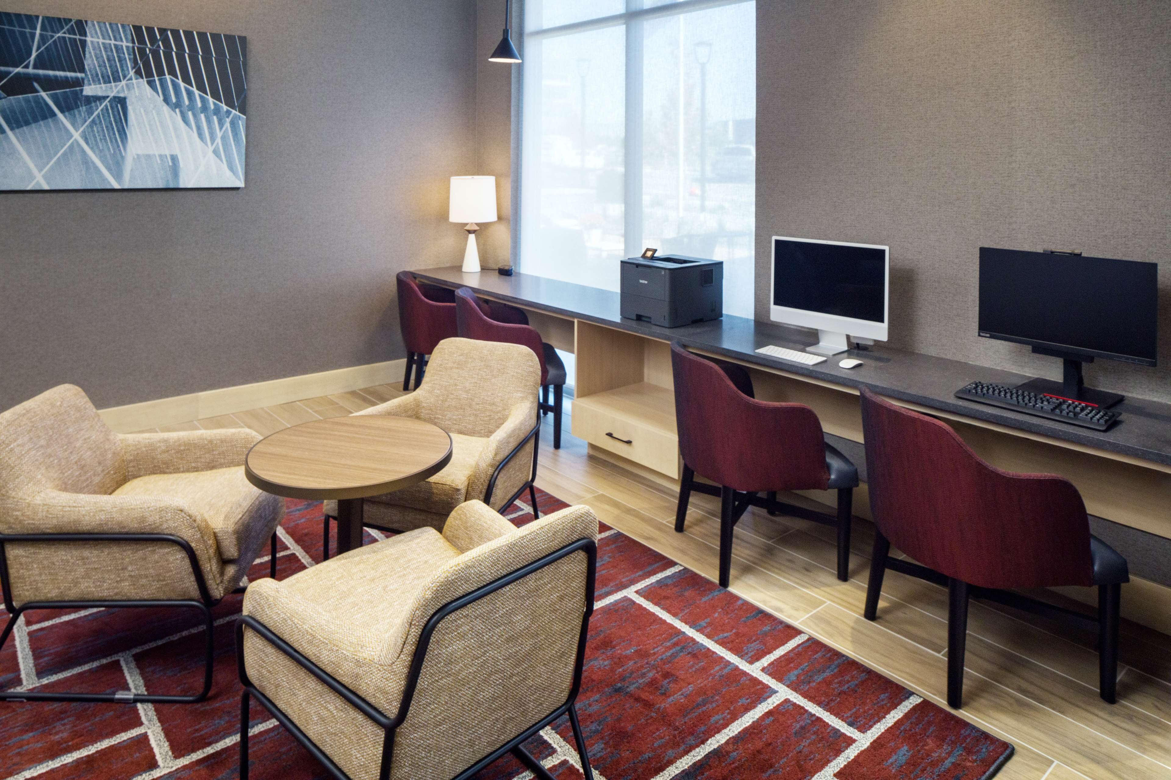A modern hotel room with a desk, two chairs, a small table, a lamp, and a computer setup.