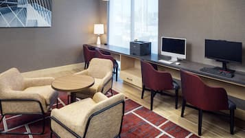 A modern hotel room with a desk, two chairs, a small table, a lamp, and a computer setup.