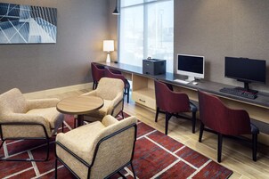 A modern hotel room with a desk, two chairs, a small table, a lamp, and a computer setup.