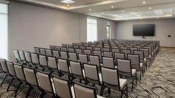 Sala de conferência com fileiras de cadeiras, uma tela grande e piso coberto de carpete.