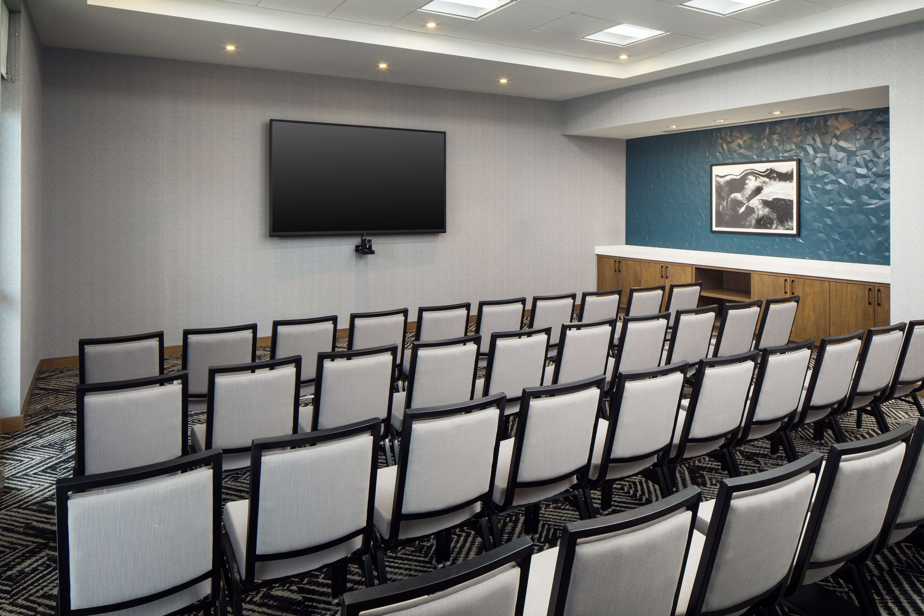Uma sala de conferências com fileiras de cadeiras, uma TV de tela plana grande, um balcão de madeira e um painel decorativo.
