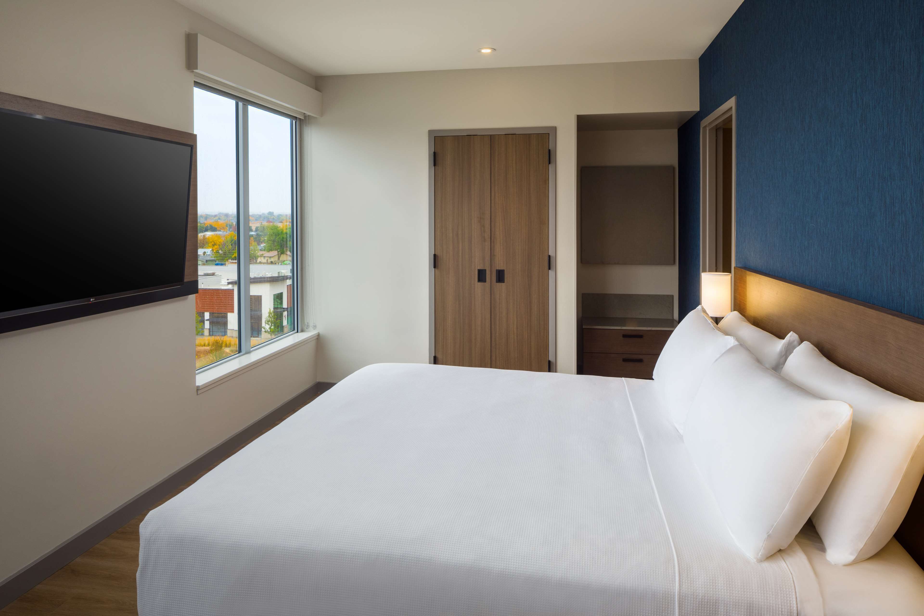 A modern hotel room with a large bed, a TV mounted on the wall, a window with a view of buildings, and a wooden wardrobe.