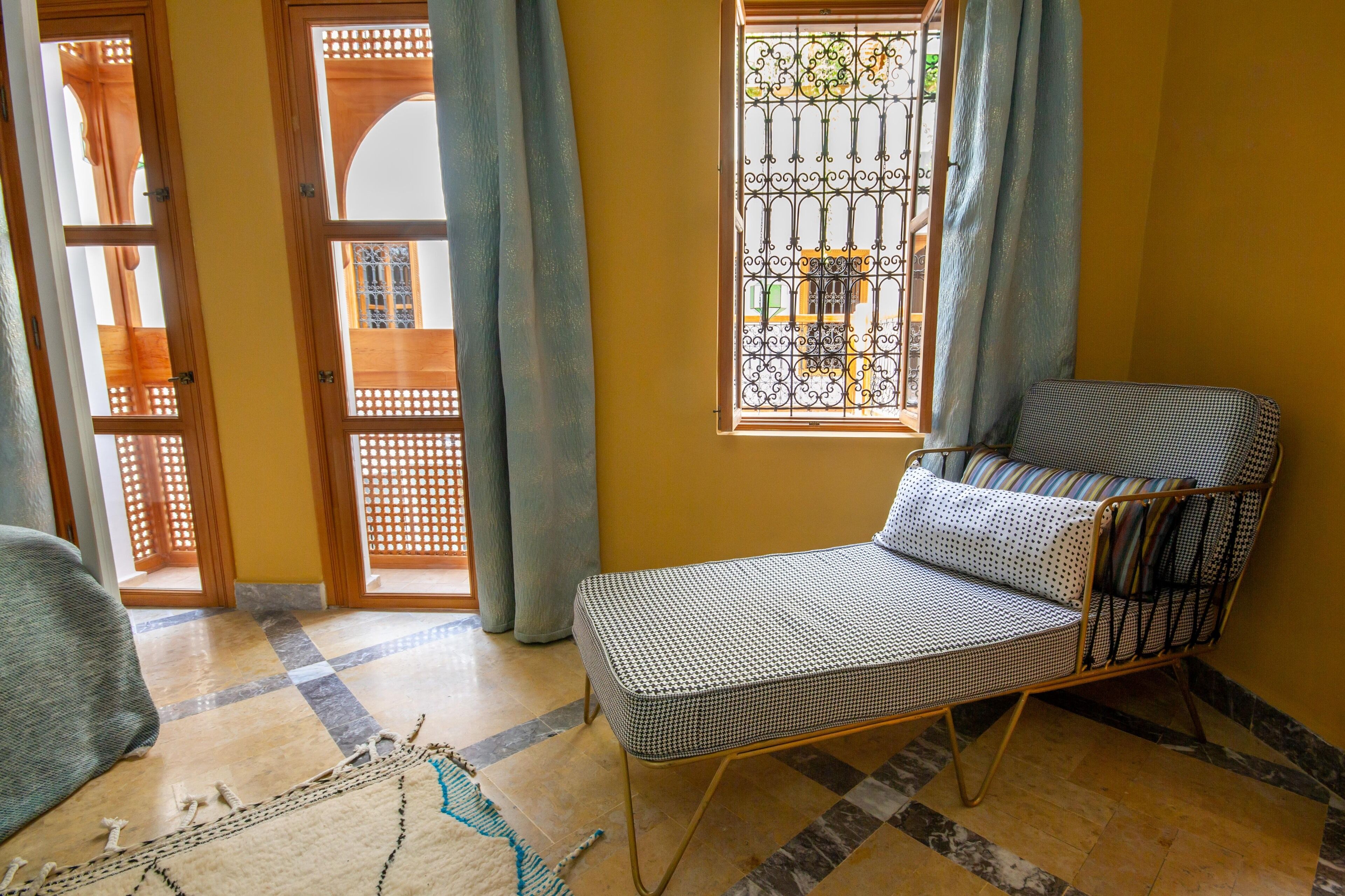 A room with a patterned sofa, a patterned rug, a window with decorative iron bars, and a view of a building outside.
