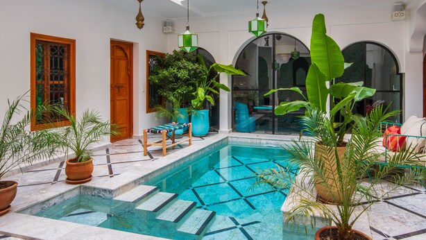 A pool area with steps leading into the water, surrounded by potted plants and wooden furniture. - Riad SofYan (Marrakech)