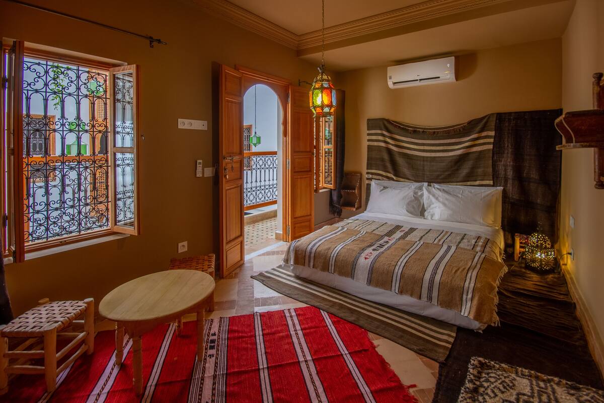A bedroom with a bed, a wooden bench, a small table, a red and white striped rug, and a decorative lantern.