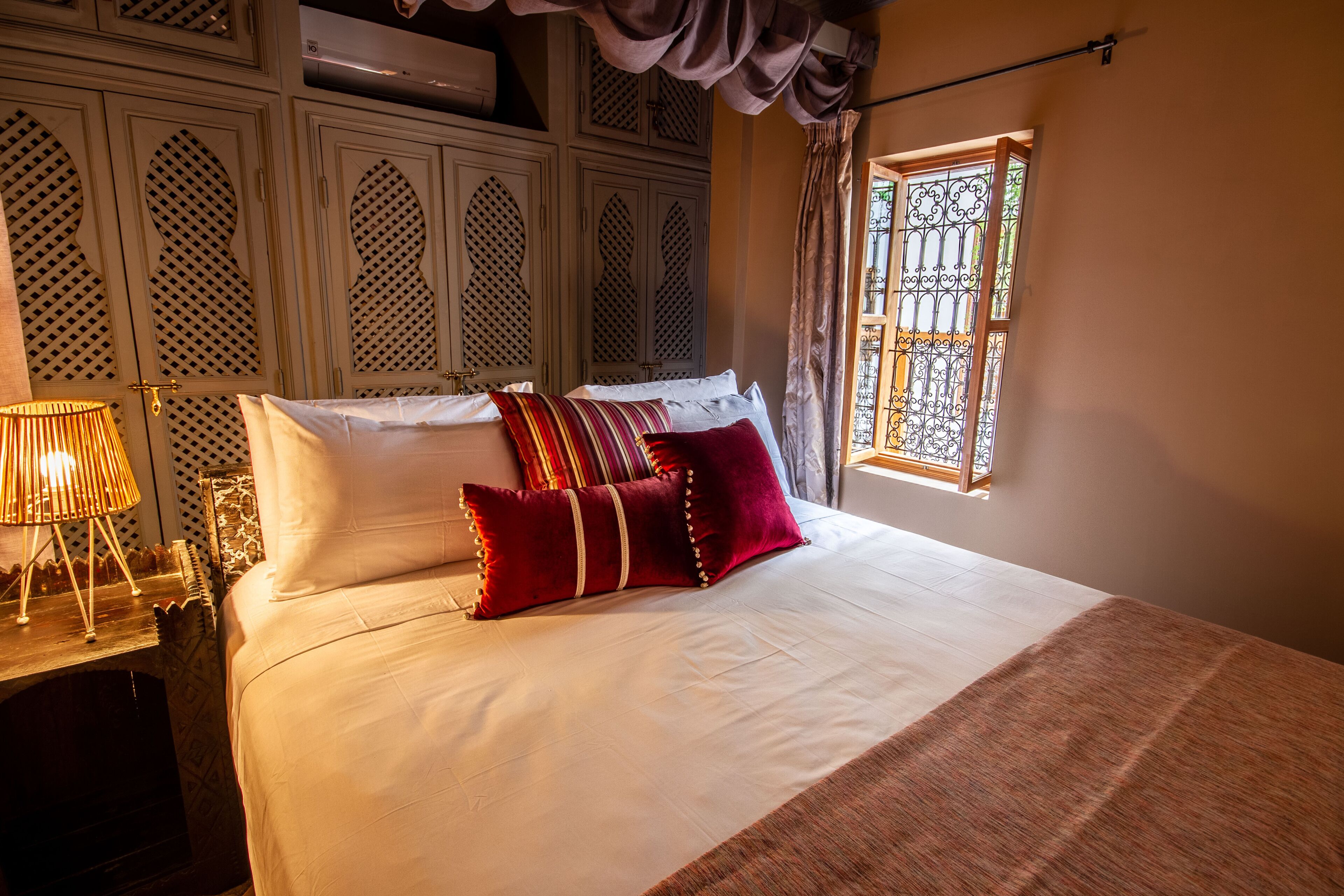 A neatly made bed with white linens and red pillows, a bedside lamp, and a window with decorative iron bars.