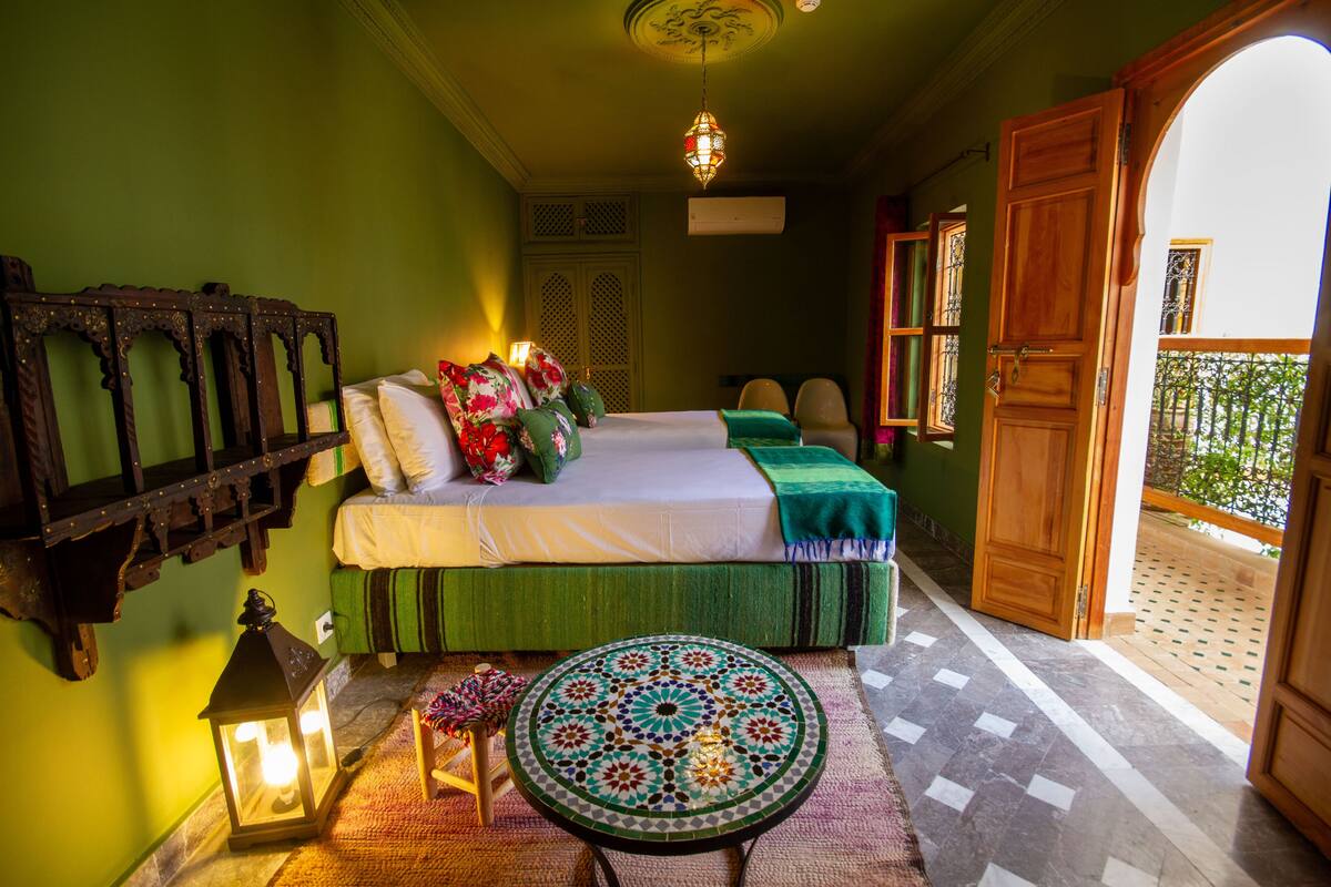 A bedroom with a bed, a round table, a hanging lantern, and a view of the outside balcony.