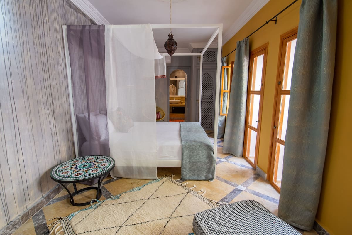 A bedroom with a bed, a small table, a bench, and a window with curtains.