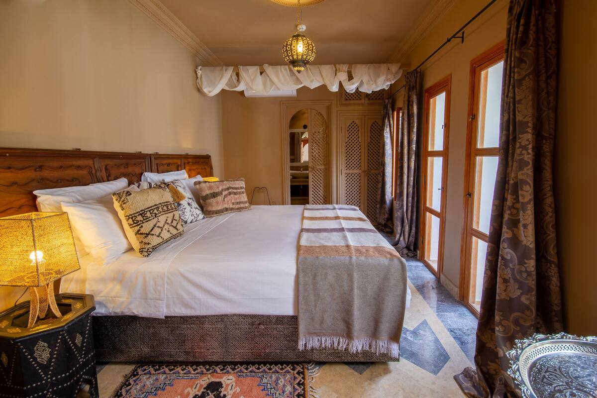 A neatly arranged bedroom with a large bed, a bedside lamp, a patterned rug, and a view of a hallway.