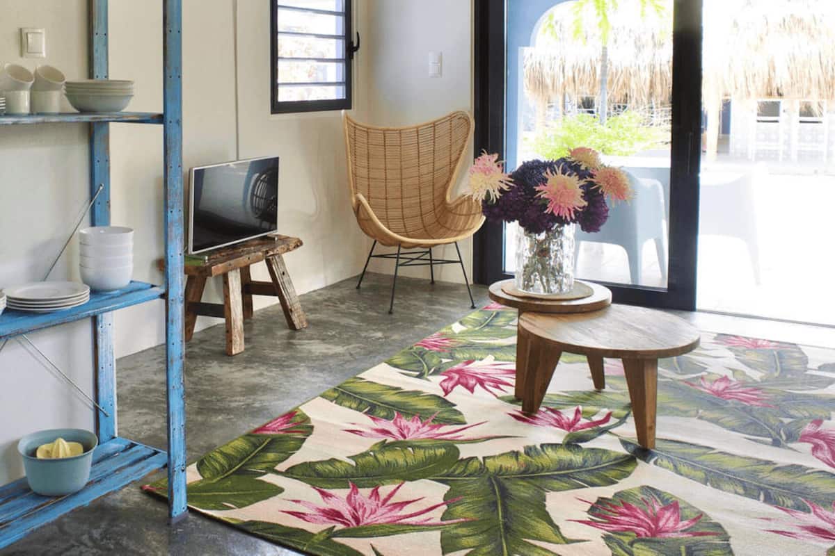 A room with a large window, a wicker chair, a round wooden table with a vase of flowers, and a colorful floral rug.