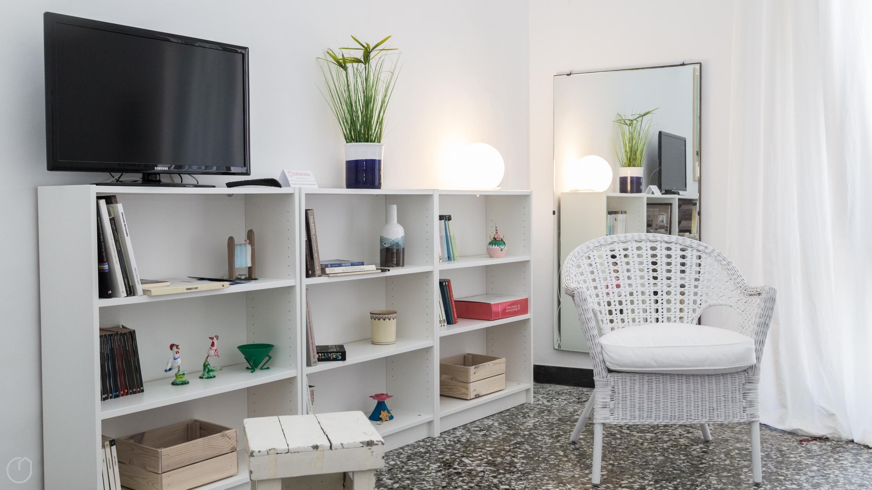 A modern living room with a flat-screen TV, white shelving units, a wicker chair, and a mirror.