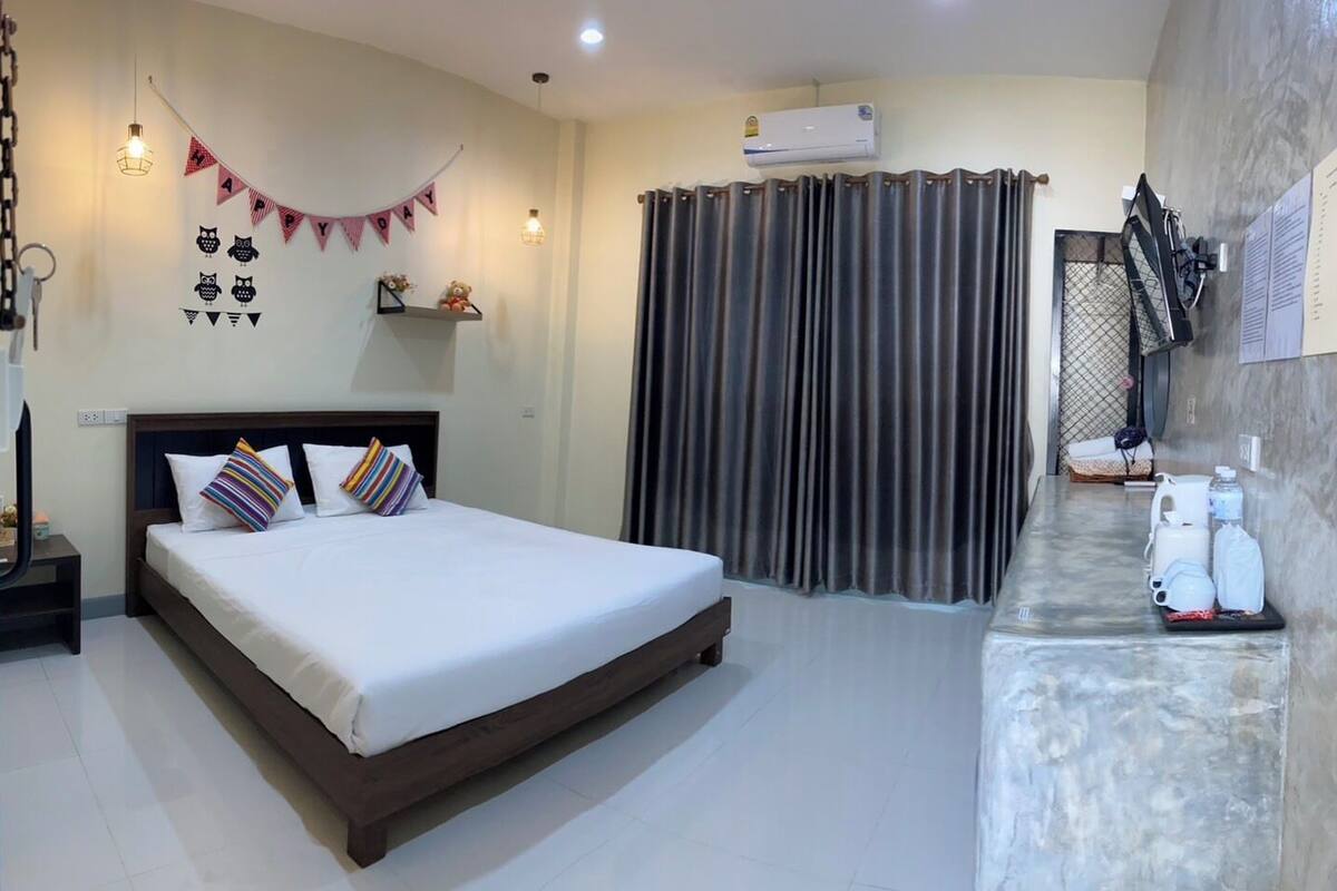 A hotel room with a bed, a bedside table, a desk with a chair, an air conditioning unit, and a curtain.