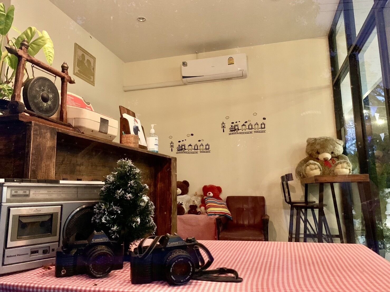 A room with a wooden shelf holding a vintage radio, a small Christmas tree, and a Canon camera. There's a teddy bear on a table and a wall-mounted air conditioner.