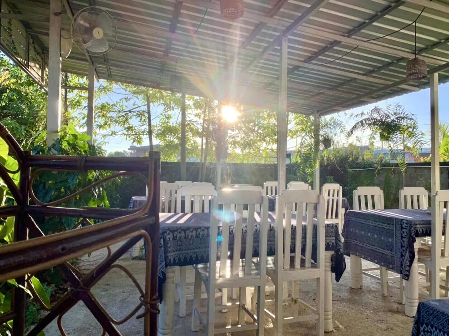 A covered outdoor seating area with white plastic chairs and tables, surrounded by greenery.