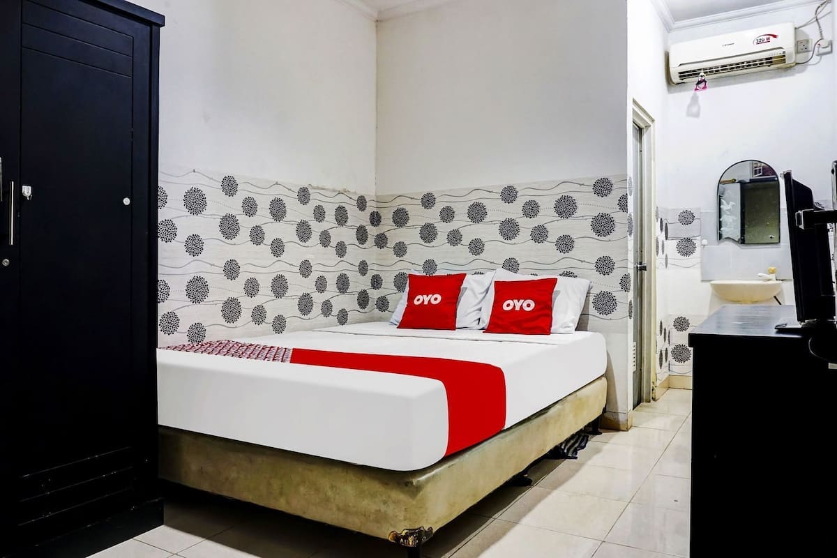 Una cama con una colcha blanca y roja, dos almohadas con la marca 'OYO', un armario negro y un baño con un lavabo y un espejo.