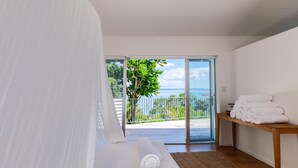 Ein Schlafzimmer mit Bett, einer Bank mit Handtüchern, Blick auf das Meer und einem Balkon mit einem Baum.