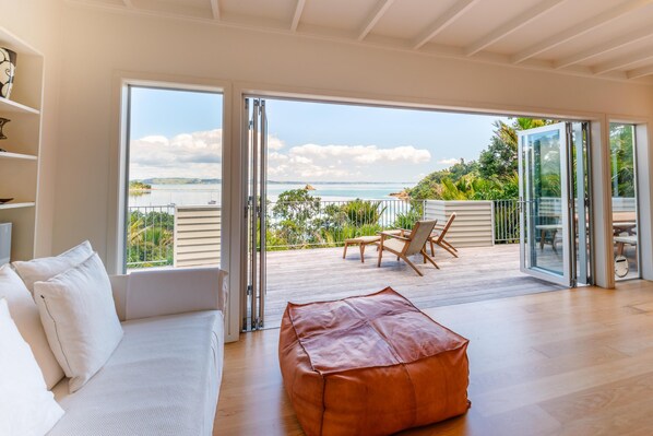 En rummelig stue med en stor skydedør, der fører ud til en balkon med havudsigt. Der er en hvid sofa og en læderpuff i værelset.