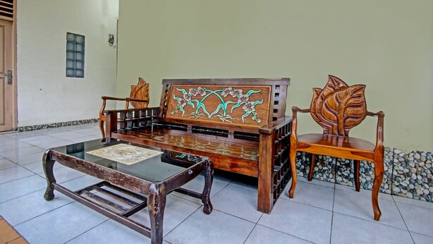 A traditional wooden sofa with intricate floral carvings, a matching wooden chair, and a glass-top coffee table in a well-lit room with tiled flooring. - OYO Life 91268 Homestay Bu Erma Syariah (Palembang)
