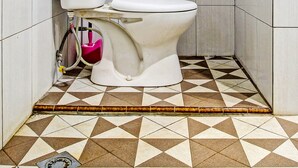 A small, tiled bathroom with a toilet, a white tank, and a pink bidet attachment.