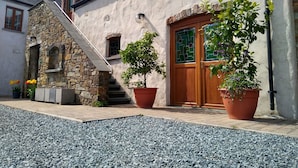 Un escalier en pierre mène à une porte en bois ornée d’un vitrail vert, le tout étant entouré de plantes en pot et d’un chemin de gravier.