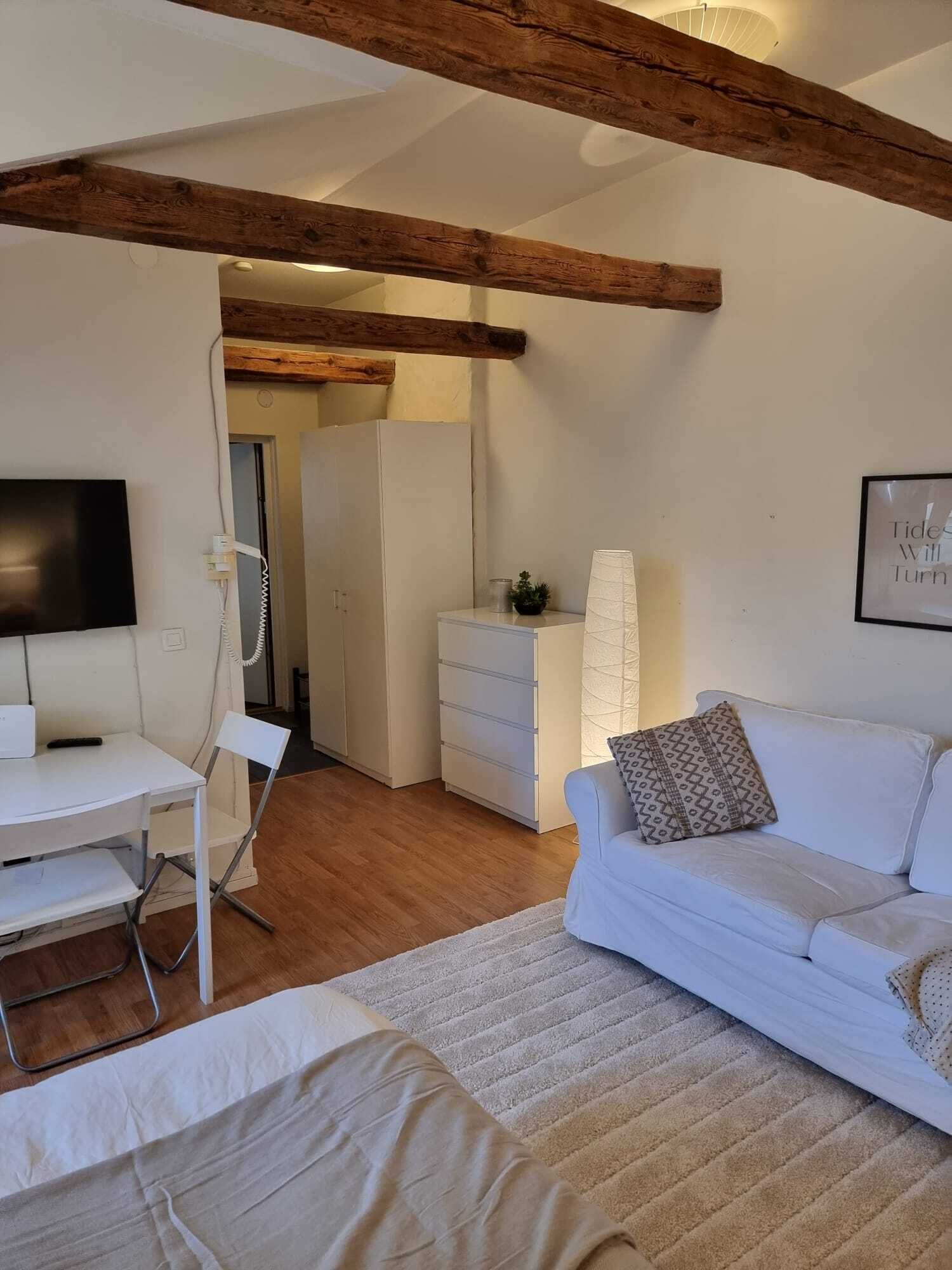 A modern living room with a white sofa, a small table, and a TV. There is a wooden ceiling beam and a white cabinet in the background.