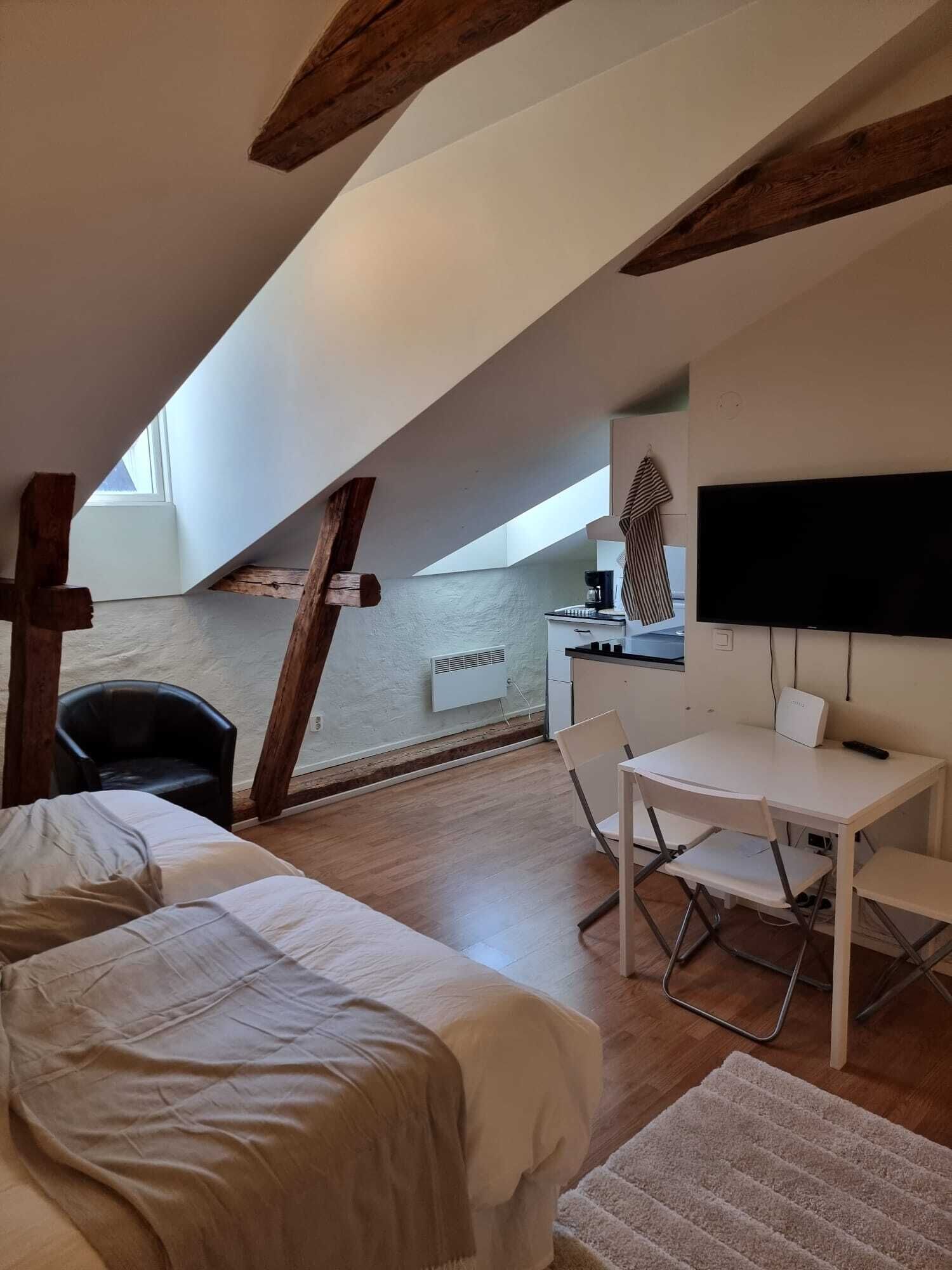 Ett litet, mysigt sovrum på vinden med ett sluttande tak, en säng, ett skrivbord med en stol, en tv och ett fönster.