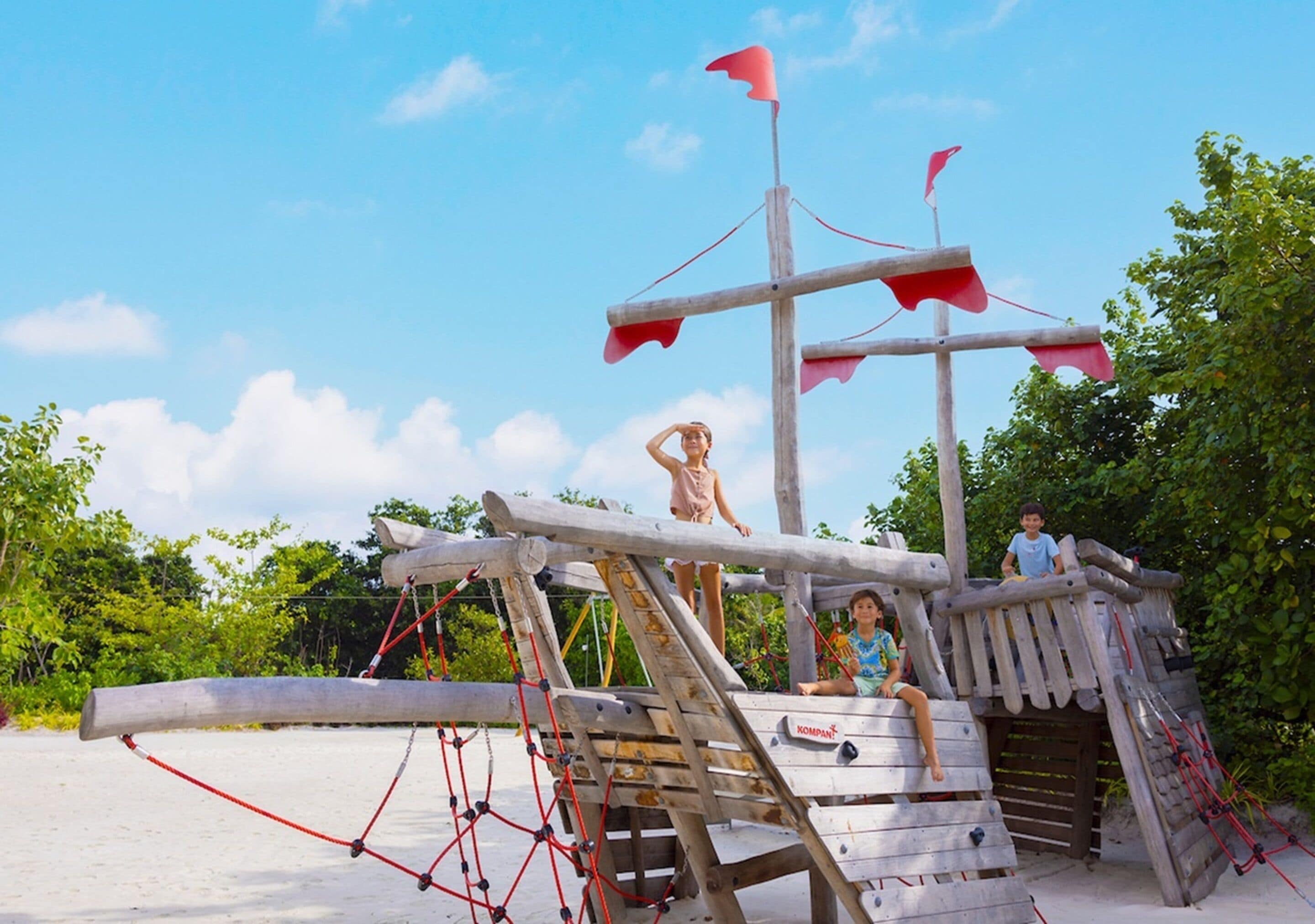 A wooden pirate ship play structure on a sandy beach with ropes and flags.