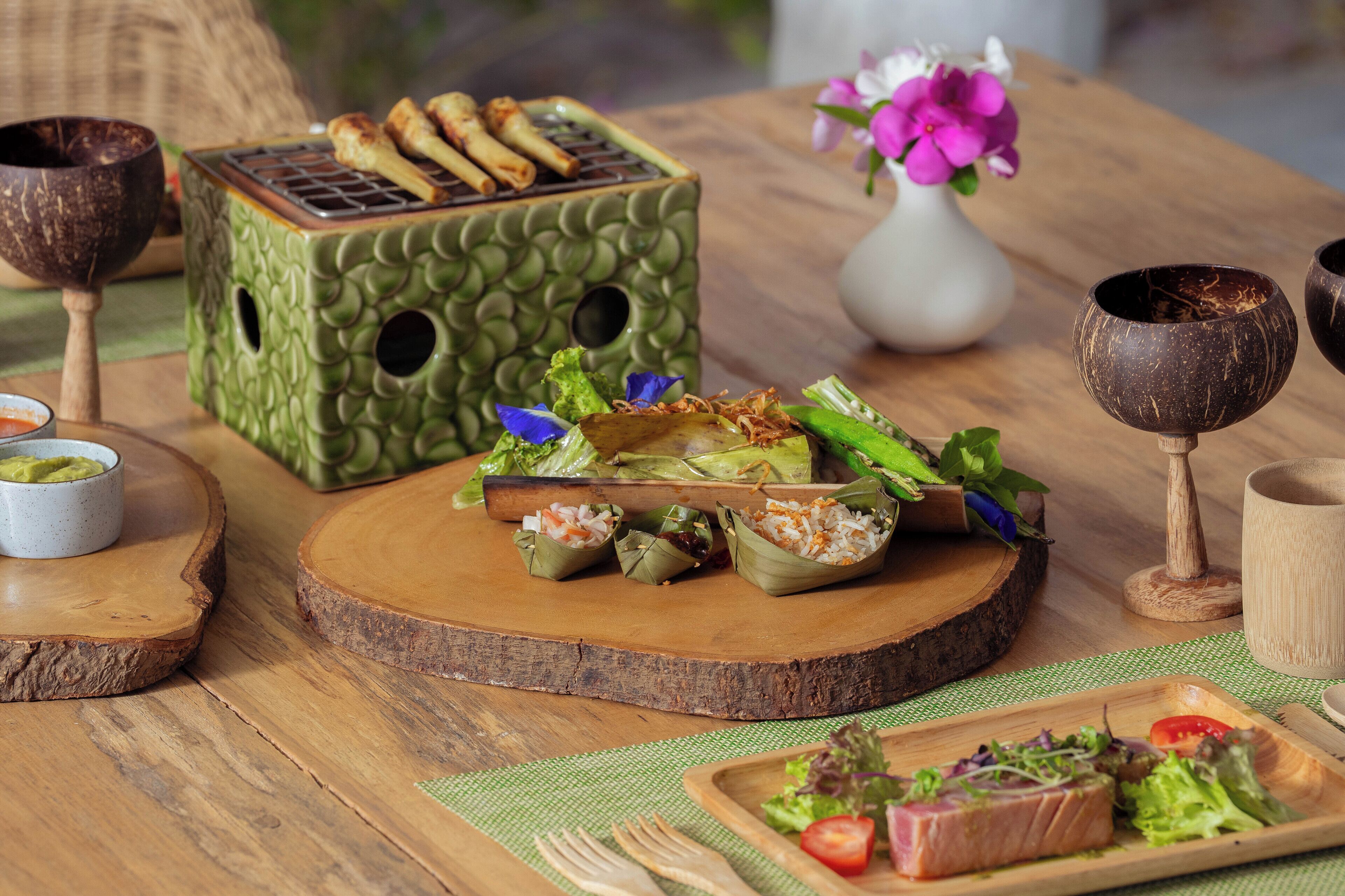 Una mesa de madera con varios platos, incluyendo una caja de cerámica verde con productos alimenticios, un florero blanco con flores y una tabla de madera con una ensalada.