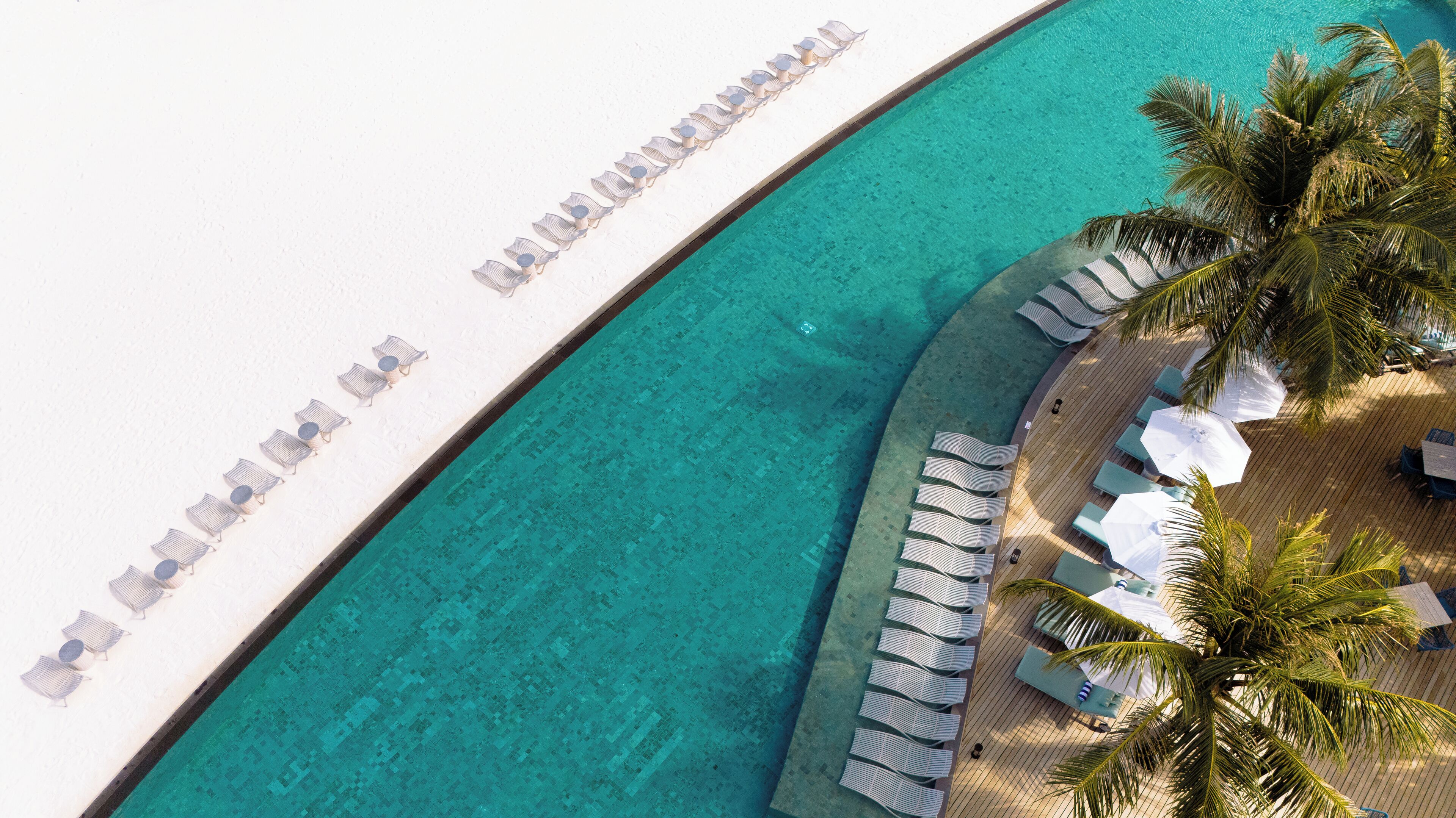 Vista aérea de um resort com piscina turquesa cristalina, praia de areia branca e espreguiçadeiras à beira da piscina.