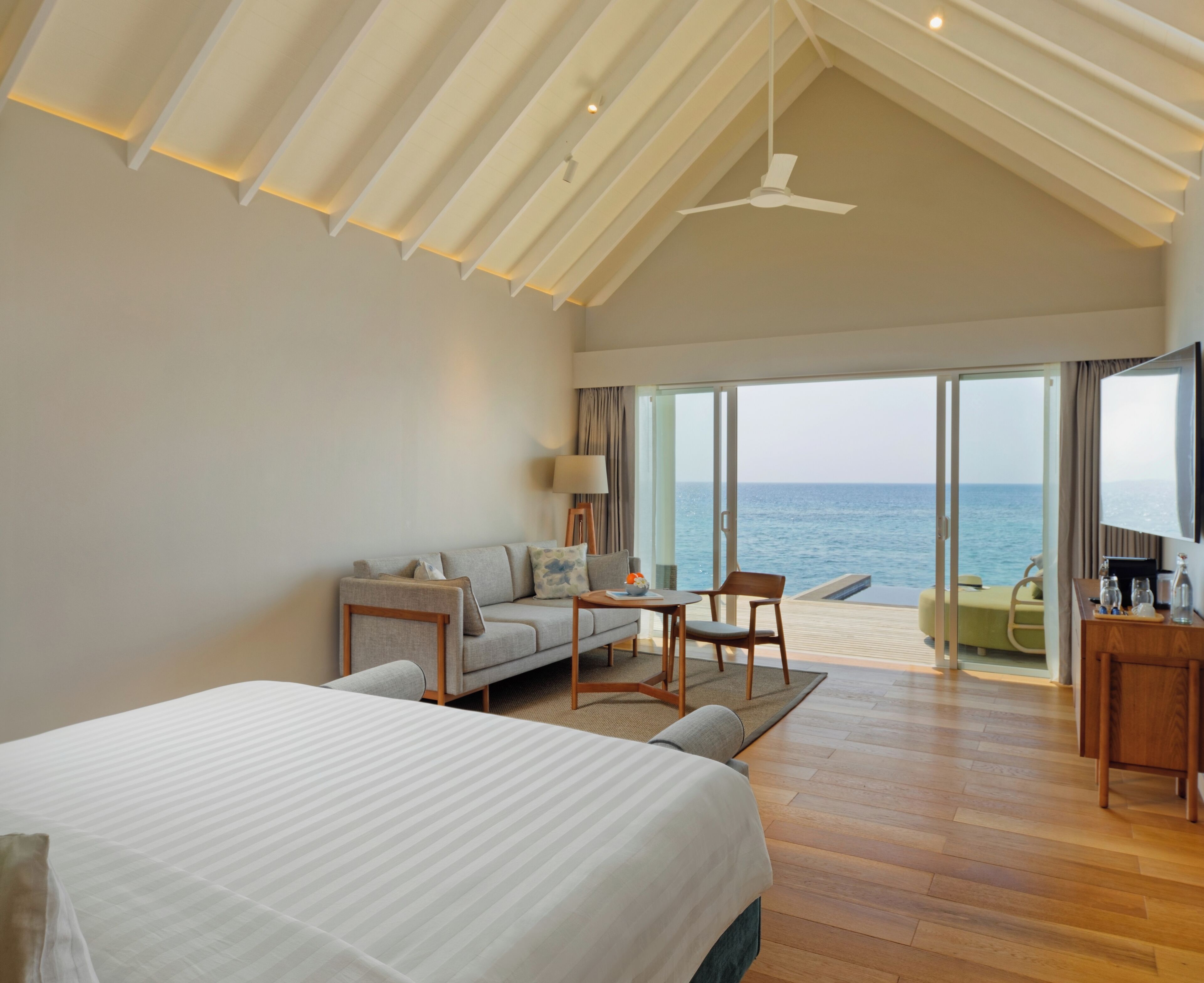 A modern hotel room with a large bed, a sofa, a dining table, and a view of the sea.
