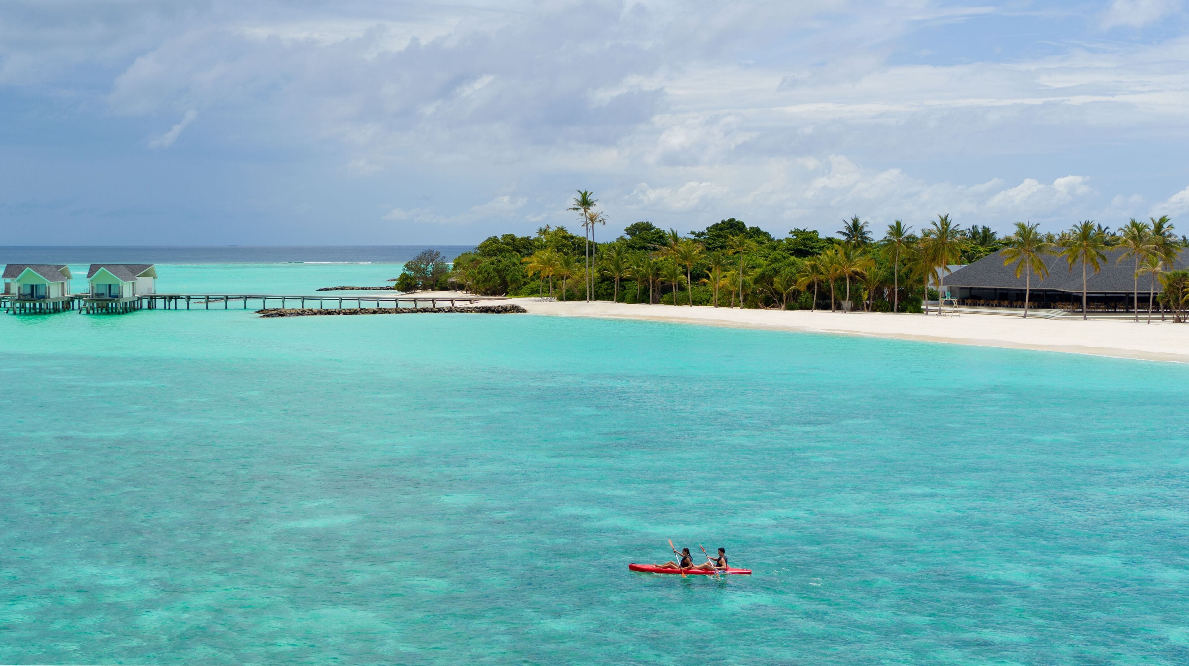 Un complejo turístico en la playa con bungalows sobre el agua, una playa de arena y palmeras.