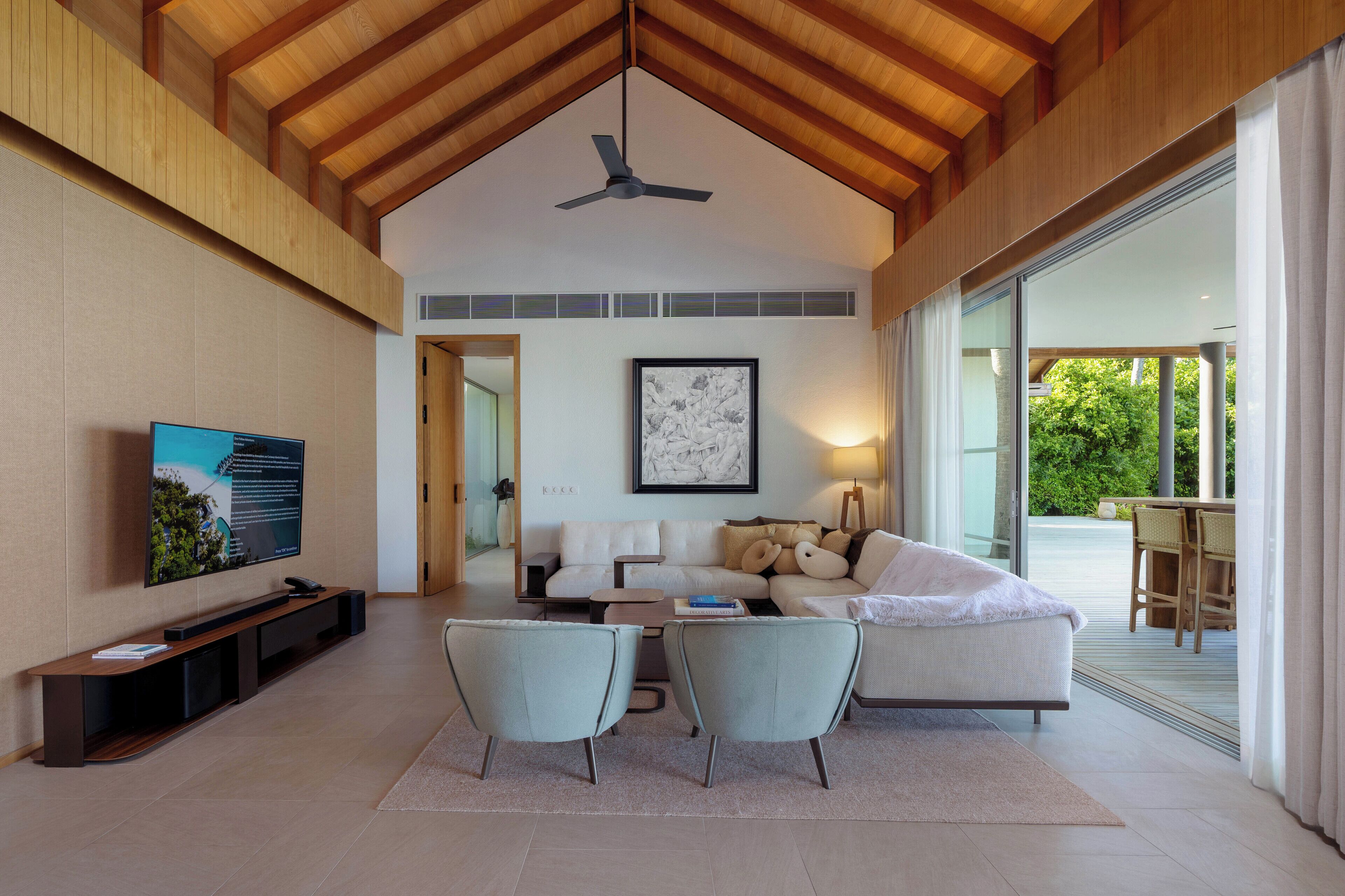 Uma sala de estar espaçosa com pé direito alto, um ventilador de teto, uma grande televisão de tela plana, um sofá, duas poltronas e um espaço de jantar com mesa e cadeiras.