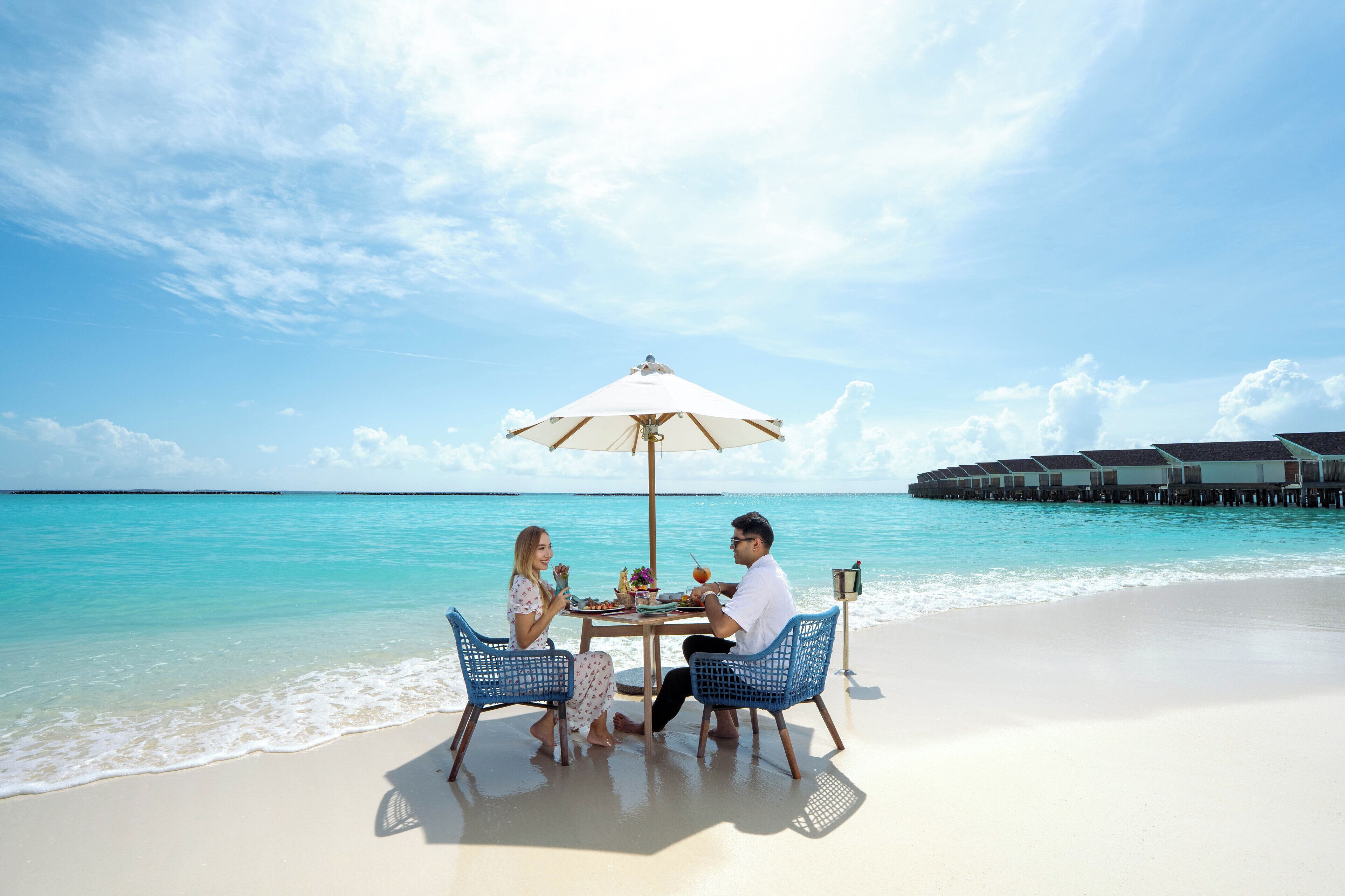 Een eettentje aan het strand met een tafel en stoelen onder een parasol, met uitzicht op turquoise water en overwater bungalows.