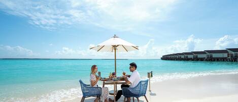 Een eettentje aan het strand met een tafel en stoelen onder een parasol, met uitzicht op turquoise water en overwater bungalows.
