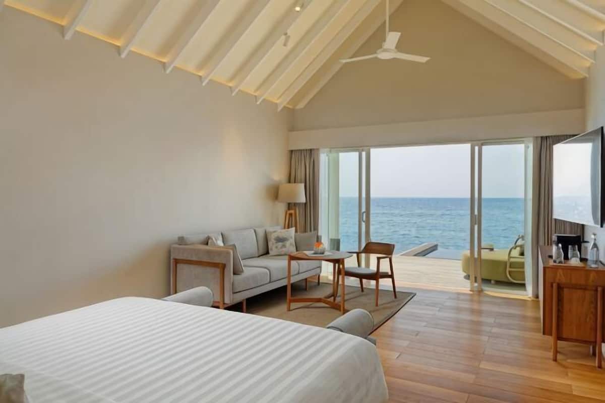 Ein modernes Hotelzimmer mit einem großen Bett, einer Couch, einem kleinen Tisch und einem Stuhl. Das Zimmer bietet Meerblick durch große Glasfenster.