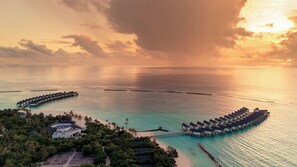 Eine Vogelperspektive auf überwasserbungalows, die durch Wege miteinander verbunden sind, umgeben von üppiger Vegetation und einem Strand.