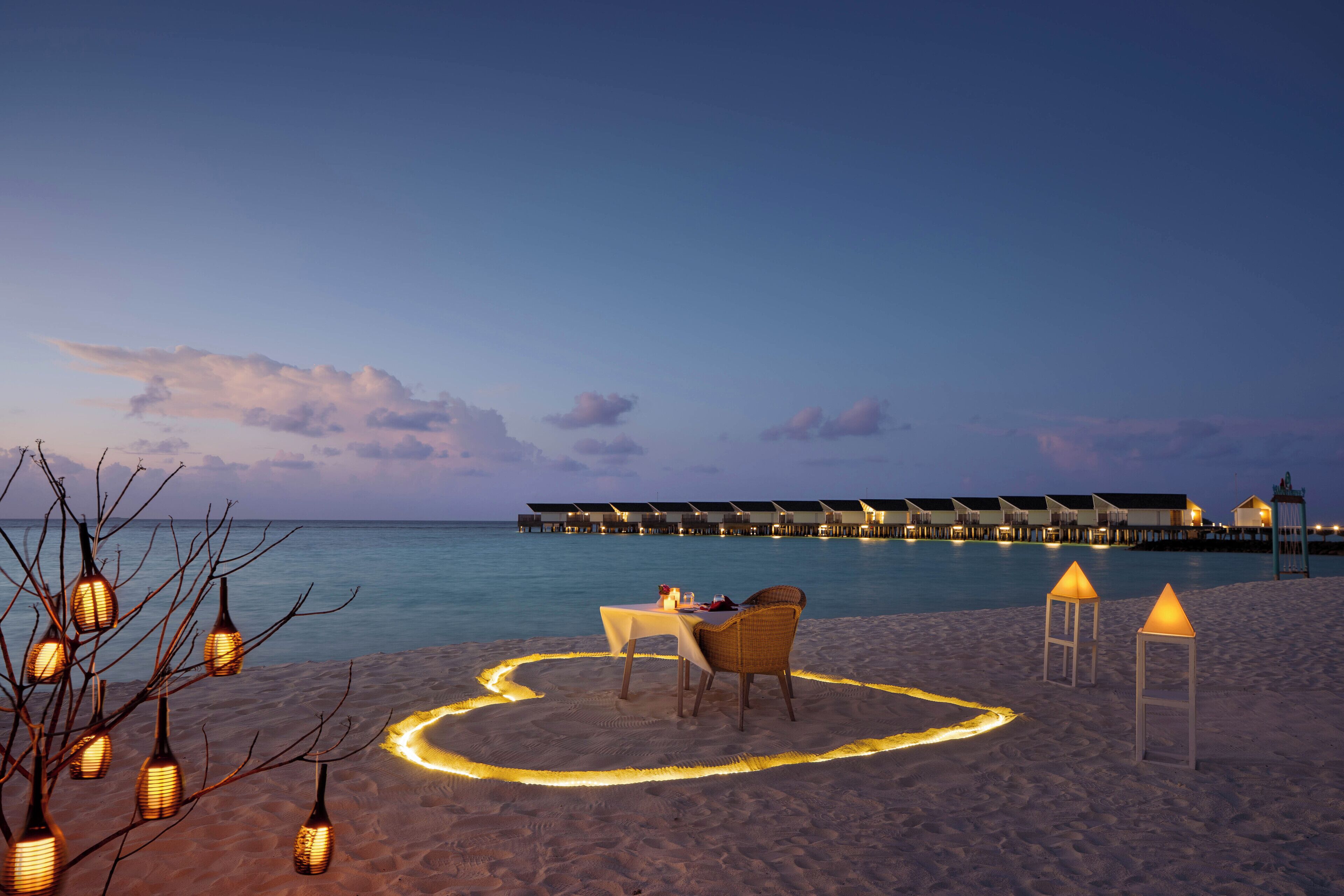 Montaje de comedor junto a la playa con una mesa y sillas, rodeado de faroles y un sendero de luces con forma de corazón en la arena, con vistas a un mar tranquilo y bungalows sobre el agua.