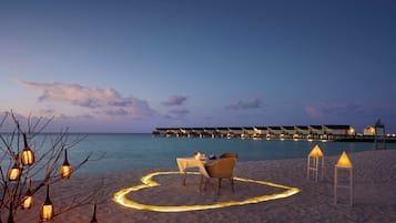 Montaje de comedor junto a la playa con una mesa y sillas, rodeado de faroles y un sendero de luces con forma de corazón en la arena, con vistas a un mar tranquilo y bungalows sobre el agua.