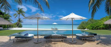 A wooden deck with lounge chairs and umbrellas overlooking a clear blue ocean and a swimming pool.