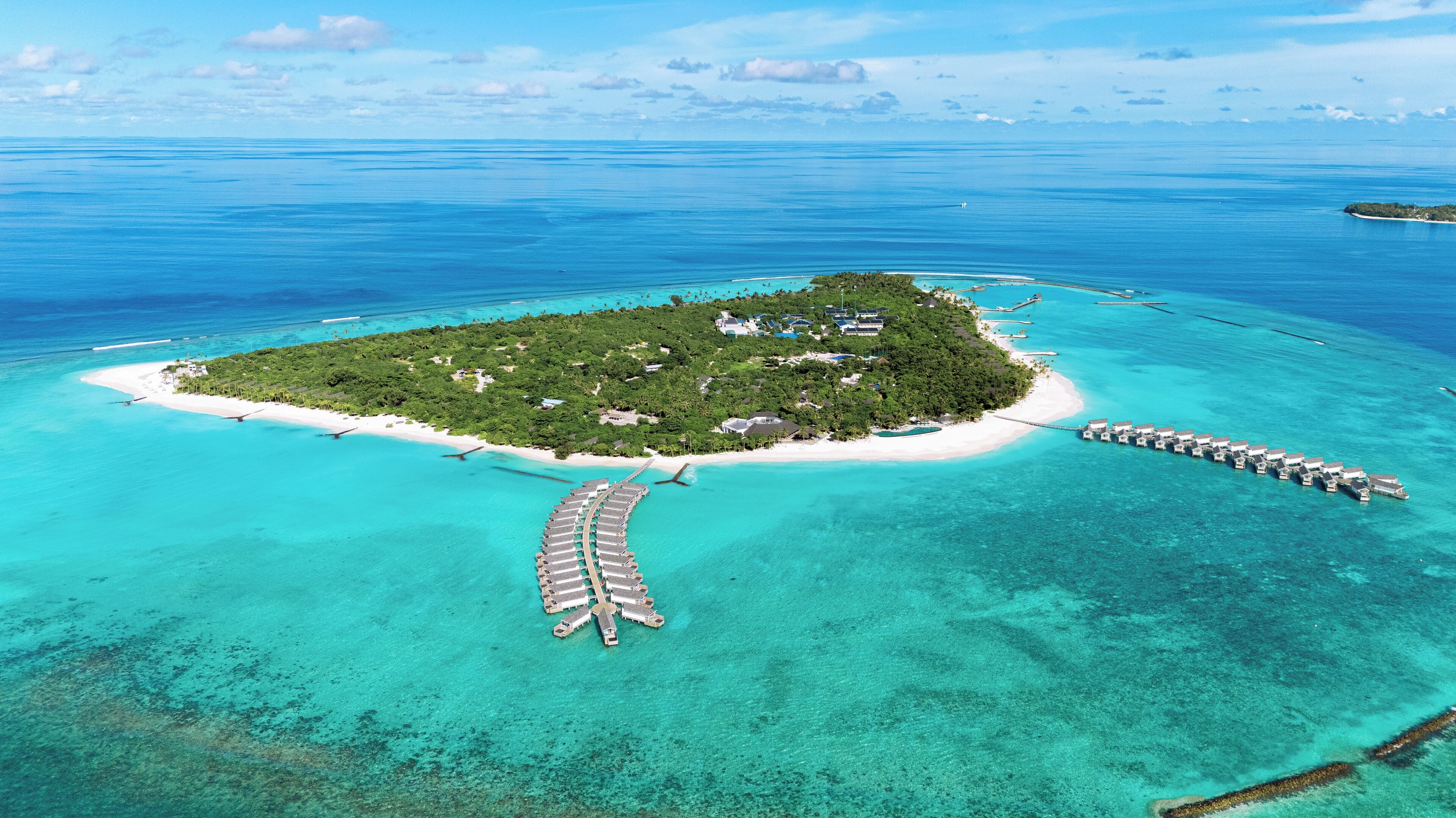 Vue aérienne d’une île tropicale aux eaux turquoise et limpides, avec une plage de sable blanc et des bungalows sur pilotis.