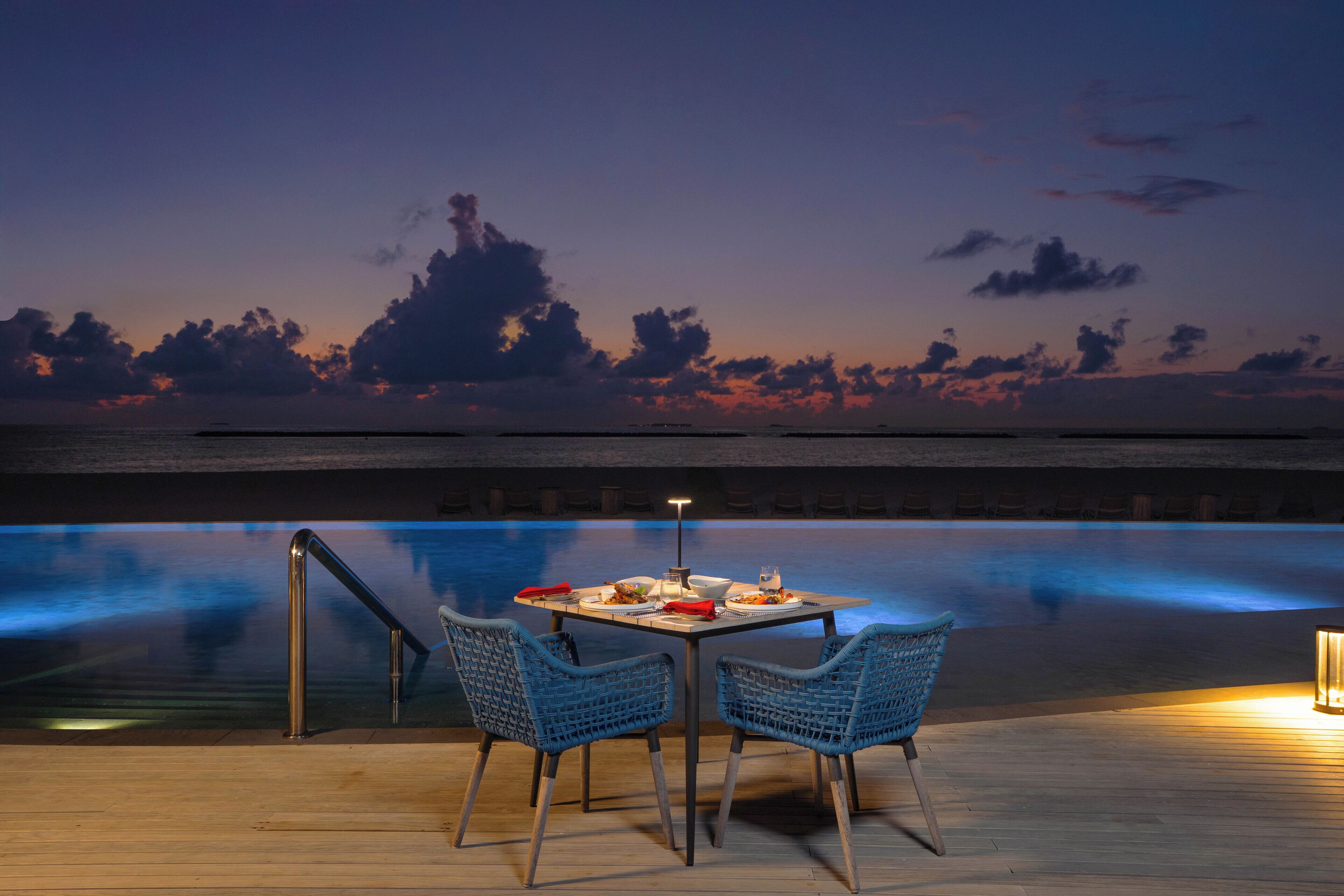 A table set for two with chairs, overlooking a pool at dusk.