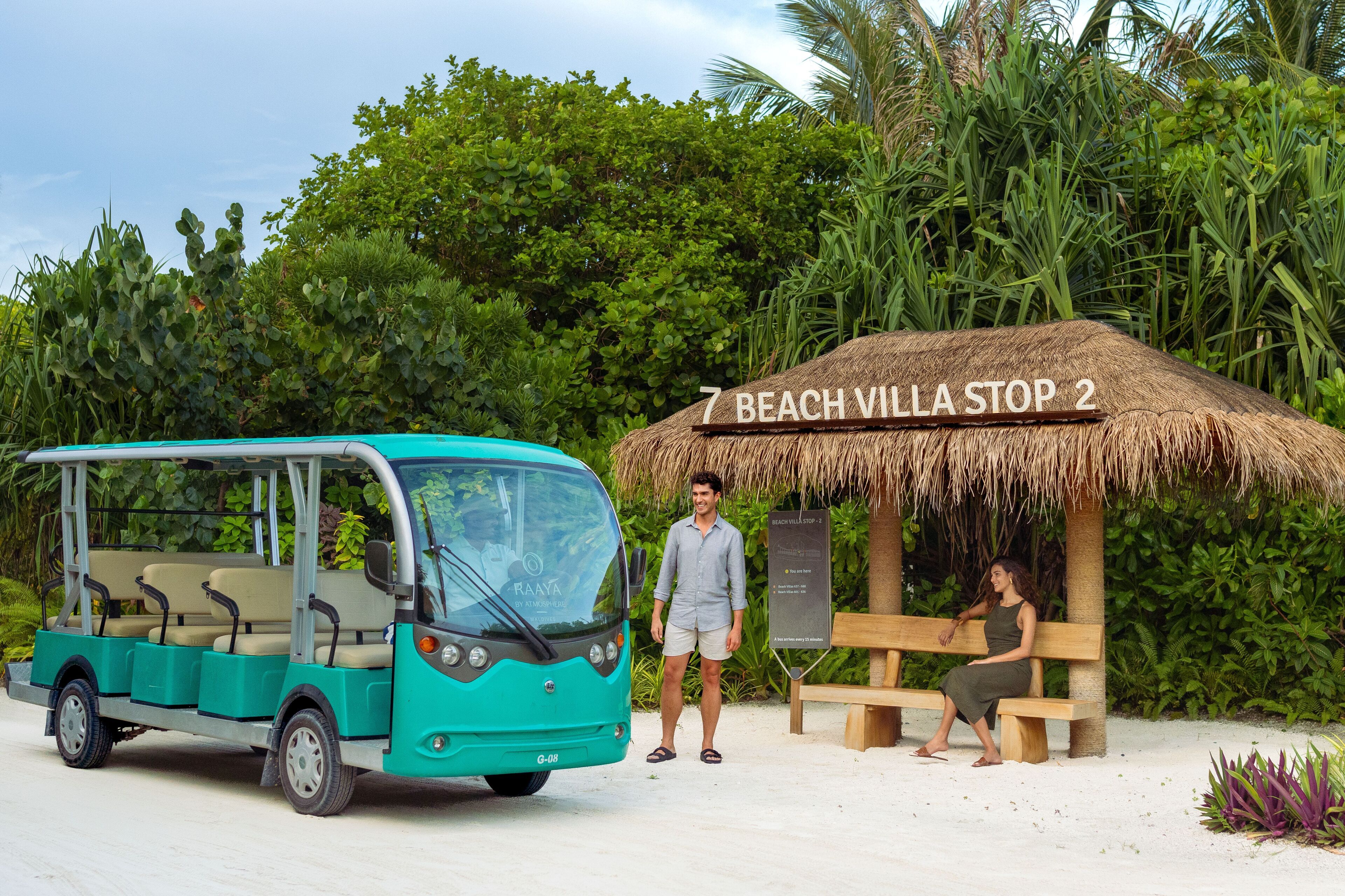 Une villa en bord de mer avec une structure au toit de chaume, un autobus turquoise et une plage de sable.