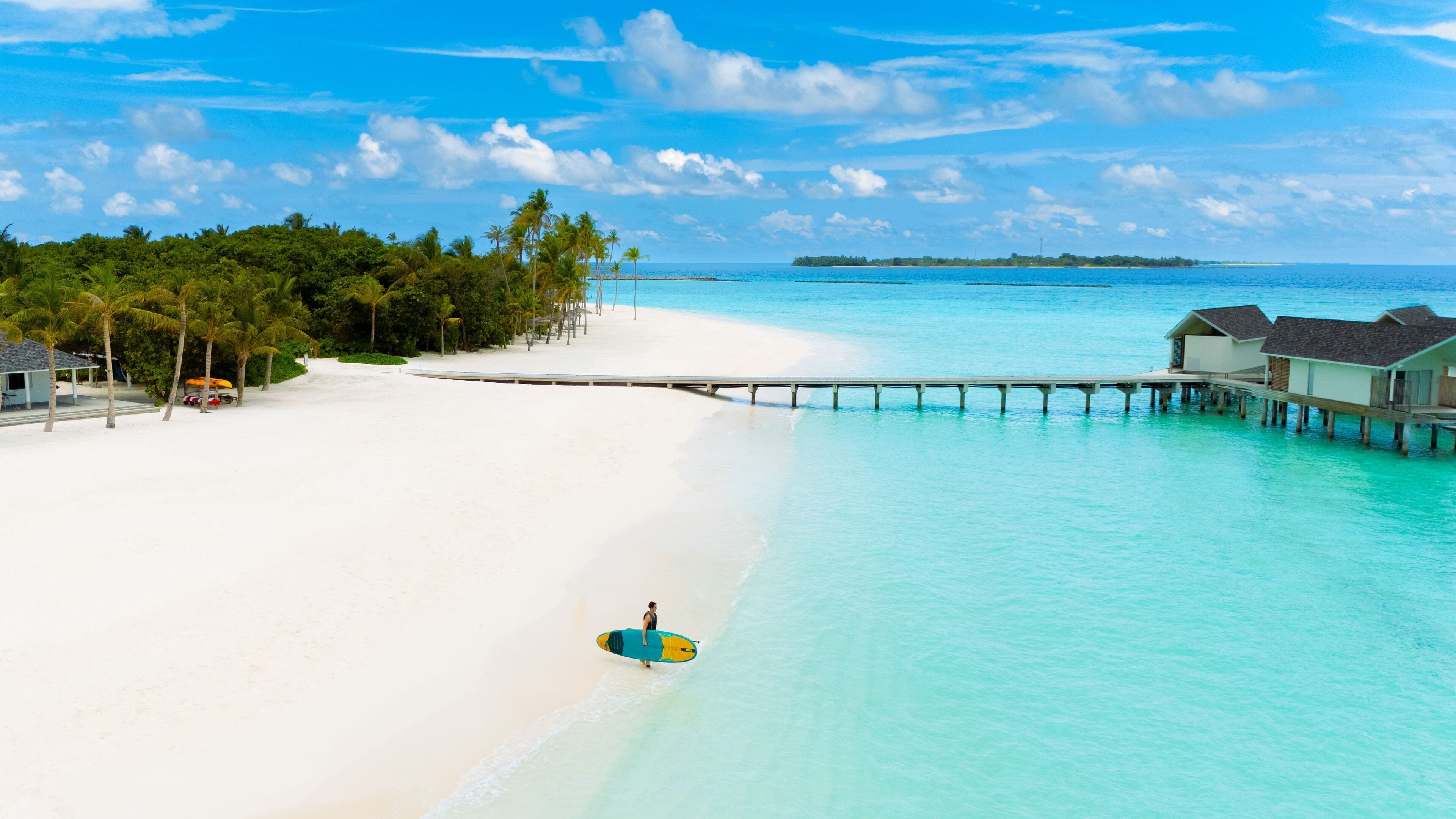 Une plage bordée d’un rivage de sable blanc, une jetée s’avançant dans une eau turquoise et des bungalows sur pilotis.