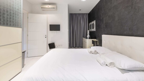 A modern hotel room with a large bed, a flat-screen TV, a vanity with a mirror, and a dark patterned wall. - Bluemarine Suite (St. Julian's)