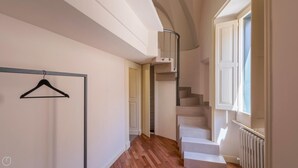 A hallway with a wooden floor, a black hanger on a metal rack, a white door, and a spiral staircase with a radiator on the wall. - Palazzo Acquaviva - Il Forno Antico (Lecce)