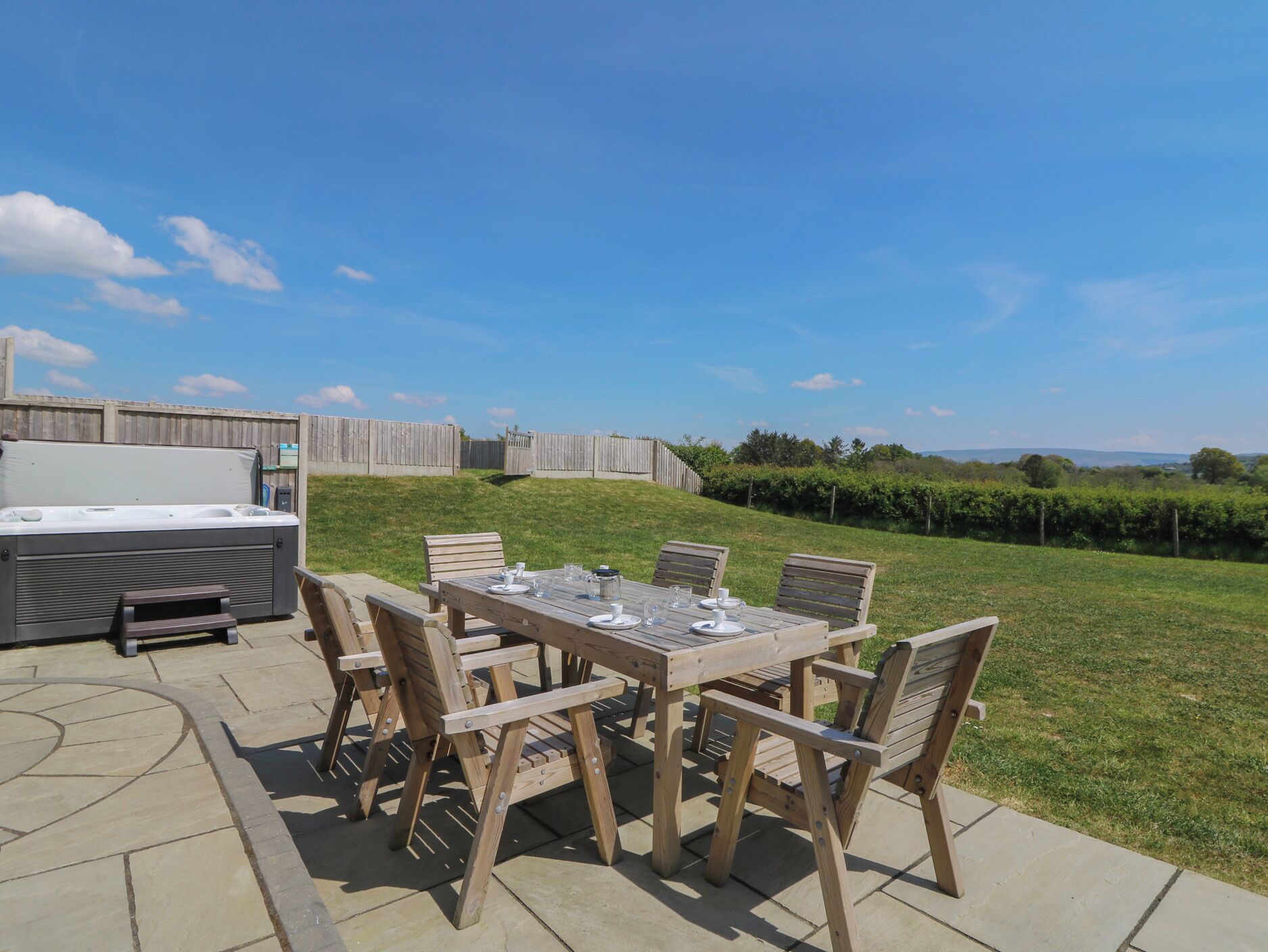 An outdoor patio with a wooden table and chairs, a hot tub, and a view of a vineyard.