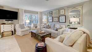 A living room with a flat-screen TV, two white sofas, a coffee table, and a bookshelf. - Gorgeous 2nd Floor Sailmaker Villa | Private Patio Overlooking Pool | Purchasable Summer Beach Club Access (Fernandina Beach)