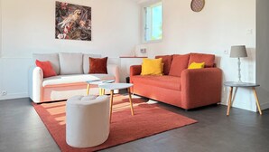 A modern living room with two sofas, a coffee table, a rug, a pendant light, and a wall clock. - La Renaissance *** - Collection La 21ème Planche (Bayeux)